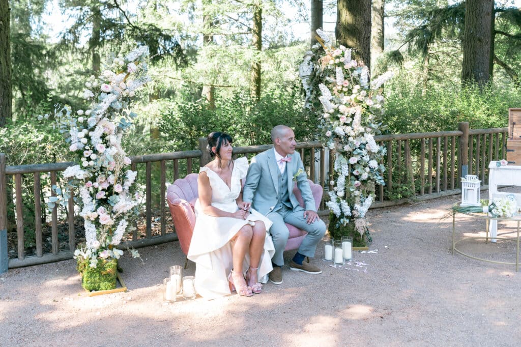 Mariage champêtre : Mariés sur canapé rose en forêt Mariés sur canapé rose entre arches fleuries, cérémonie de mariage champêtre en forêt.