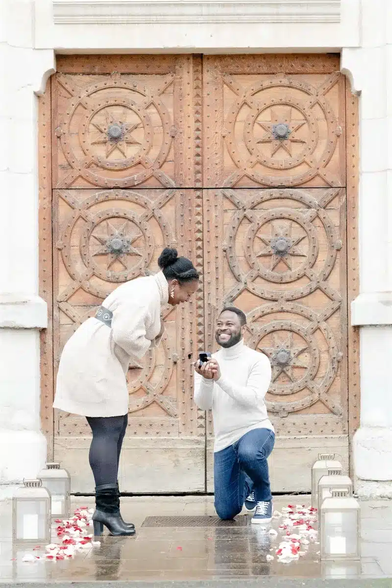 Un homme agenouillé avec une bague devant une femme souriante, une grande porte sculptée en bois en arrière-plan.