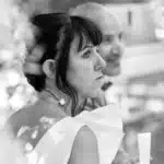 Portrait noir et blanc d'une femme pensant, vêtue d'une robe blanche à volants, avec un homme souriant flou en arrière-plan.