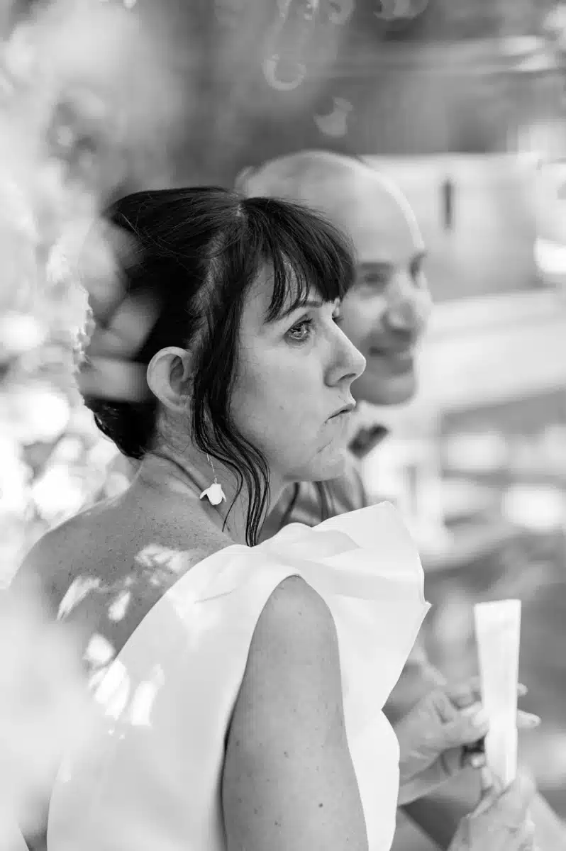 Portrait noir et blanc d'une femme pensant, vêtue d'une robe blanche à volants, avec un homme souriant flou en arrière-plan.