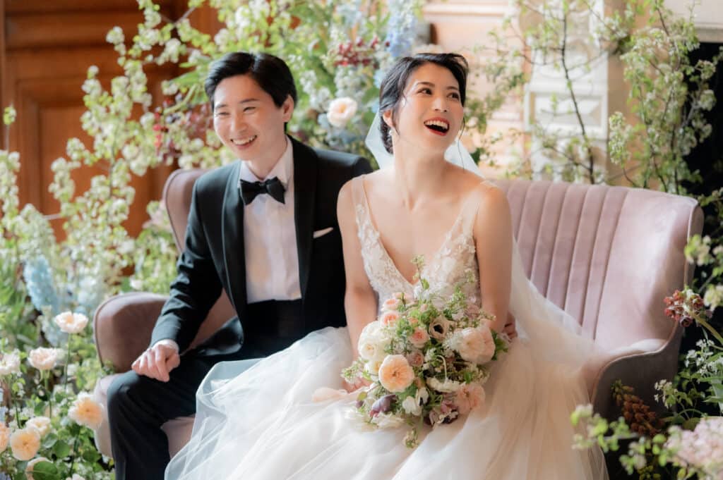 Mariage Élégant : Couple Heureux sur Canapé de Fleurs Couple de mariés souriants sur un canapé rose parmi des fleurs printanières, mariée tenant un bouquet.