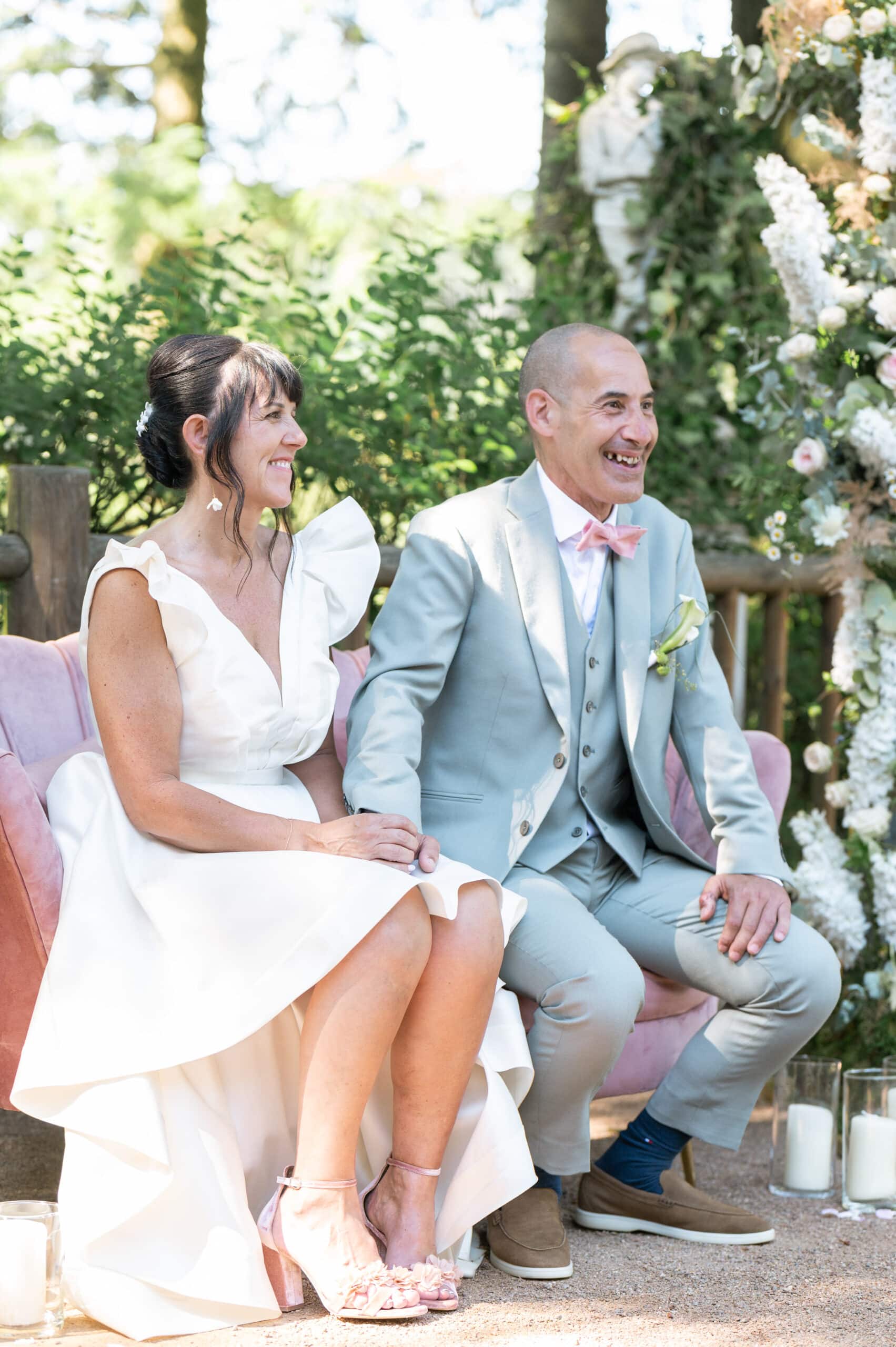 Mariés souriants assis sur un canapé rose lors d'une cérémonie en extérieur avec arche fleurie.