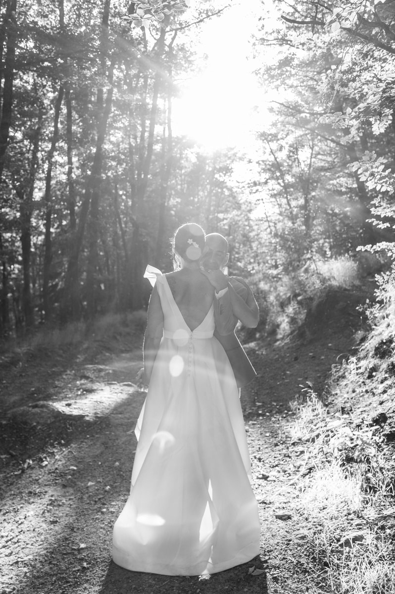 Mariés s'enlaçant sur un chemin forestier, baignés par une lumière solaire intense en noir et blanc.