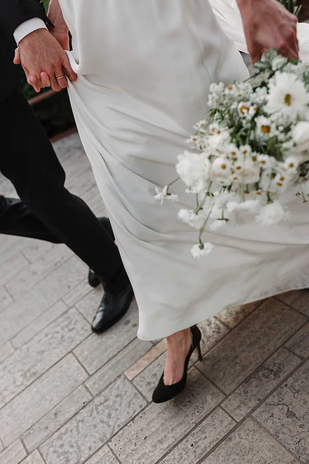 Mariés marchant main dans la main, robe en satin, bouquet de marguerites blanches et escarpins noirs sur pavés.