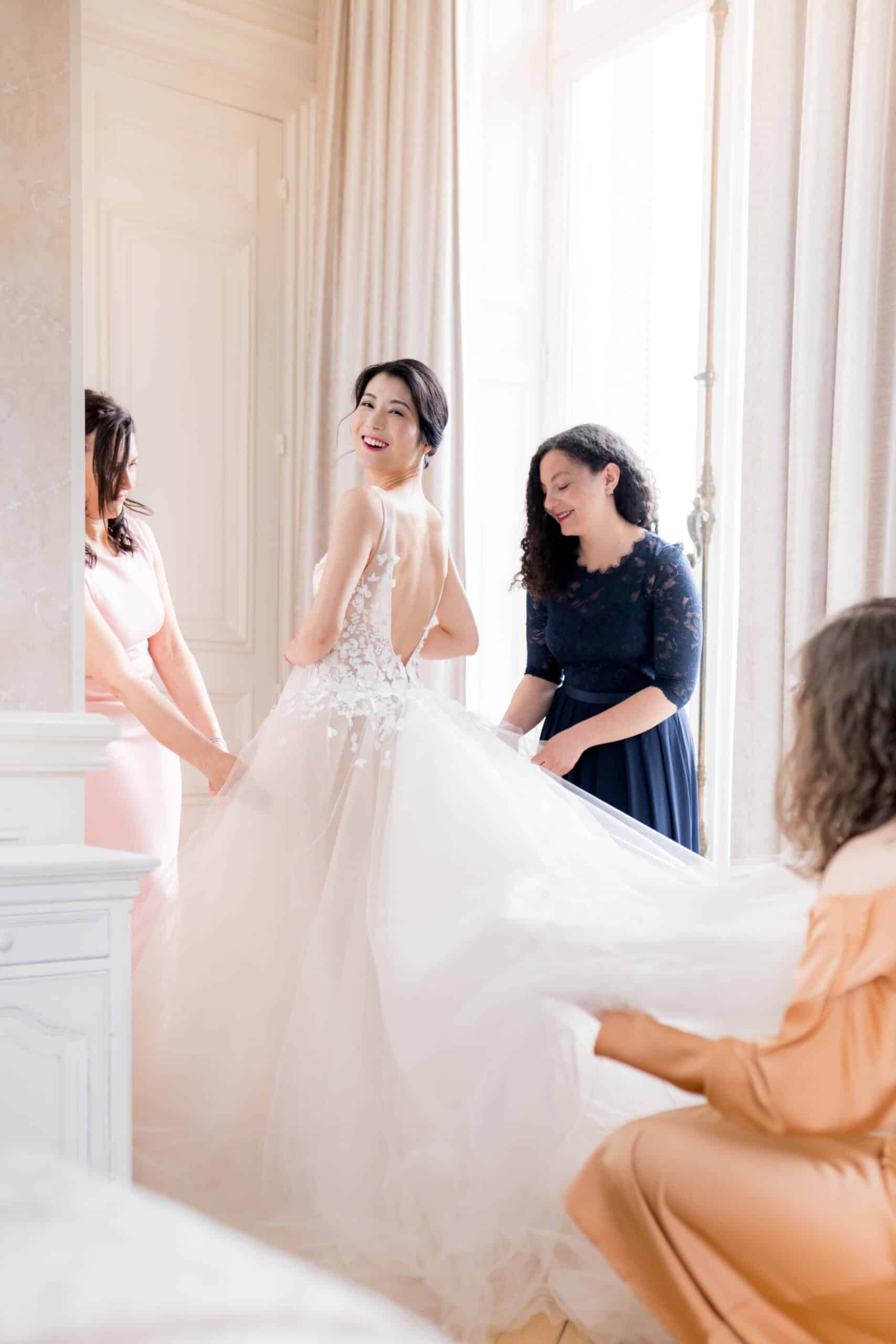Mariée souriante aidée pour sa robe fleurie par ses demoiselles d'honneur dans un intérieur élégant.