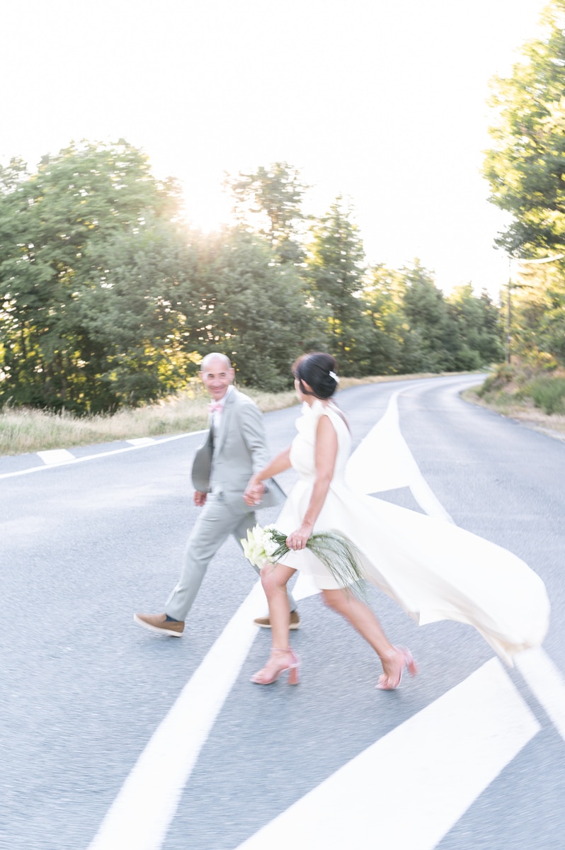 Mariés courant joyeusement sur un passage piéton en forêt, robe blanche fluide et bouquet de lys blancs.